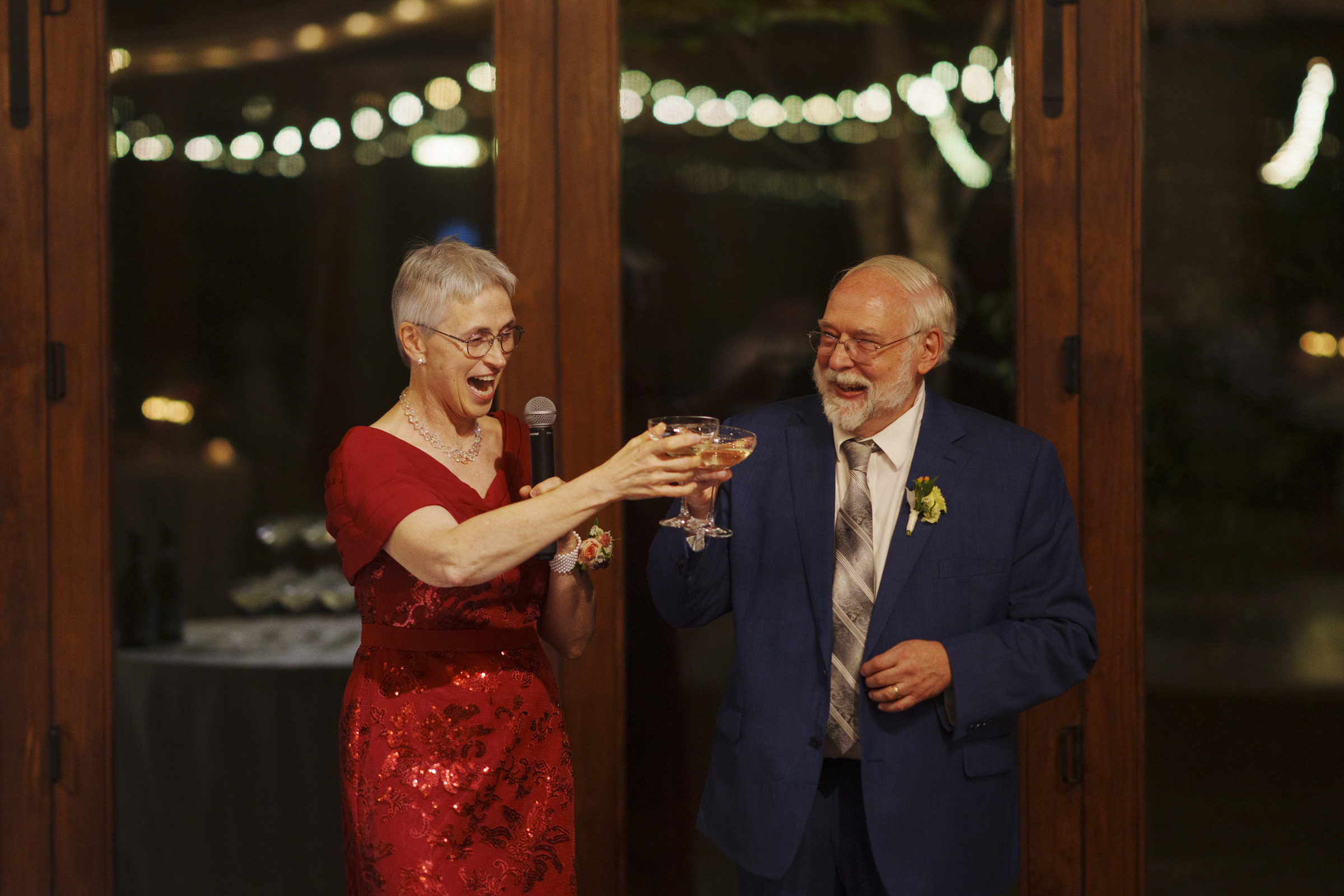 An elderly couple in formal attire is toasting with champagne glasses. The woman, in a red dress, holds a microphone and smiles at the man in a blue suit. String lights create a soft, festive background.