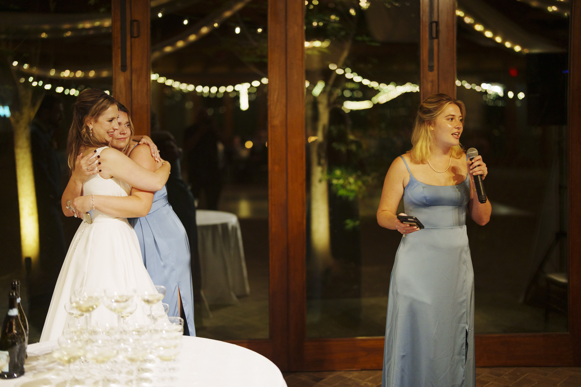 A bride in a white gown hugs a woman in a light blue dress near a wooden door with string lights above. Another woman in a similar dress stands beside them, holding a microphone and a phone. A table with stacked champagne glasses is in the foreground.