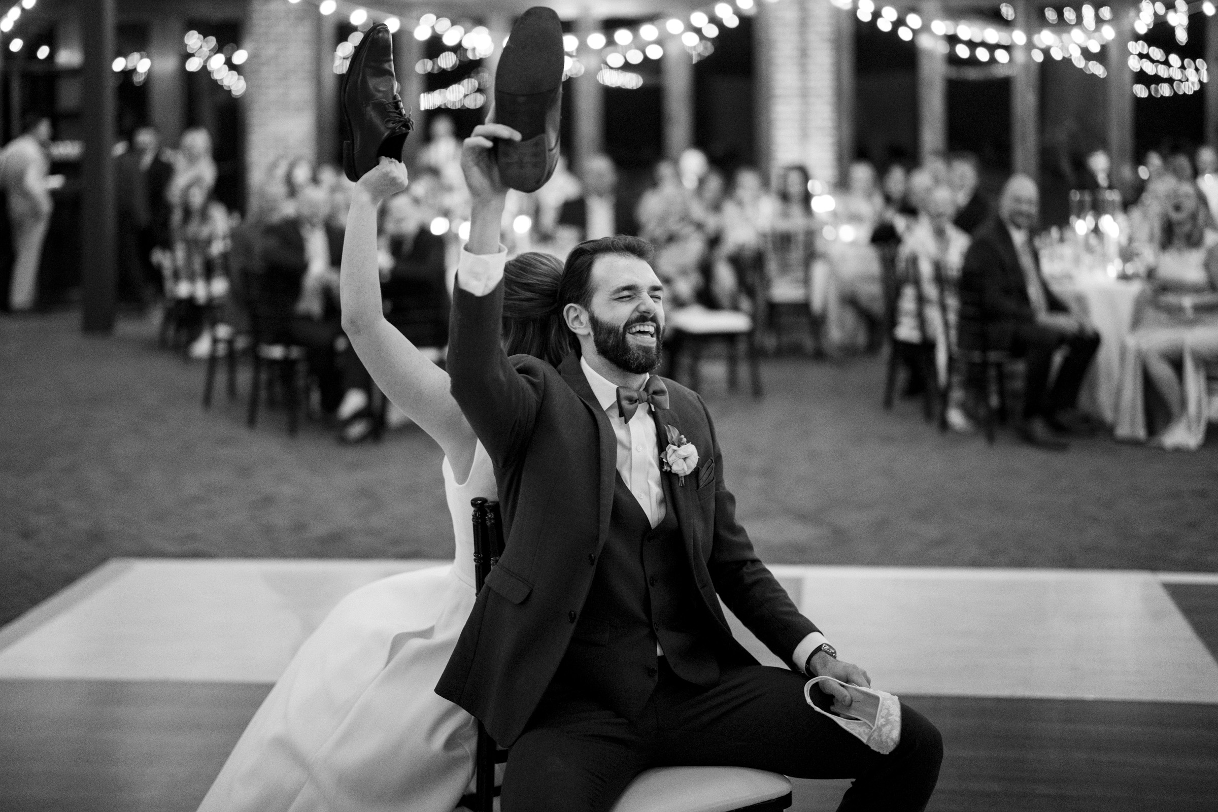 A bride and groom are seated back-to-back during a wedding game, each holding one shoe from the other. Guests are seated in the background, with string lights hanging from the ceiling, creating a festive atmosphere. The image is in black and white.