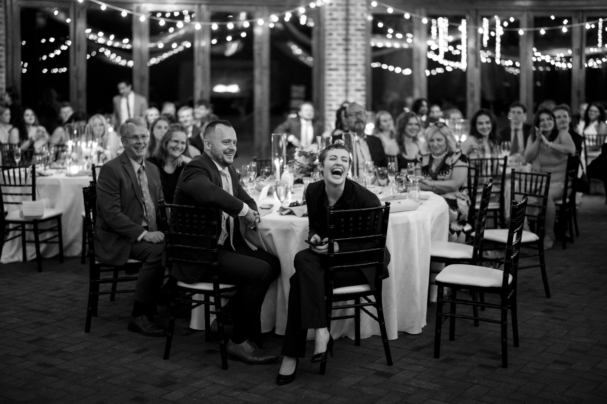 A black and white photo of a formal event with people seated at round tables, adorned with string lights. A woman in the foreground is laughing, and others are smiling, creating a lively and joyful atmosphere.