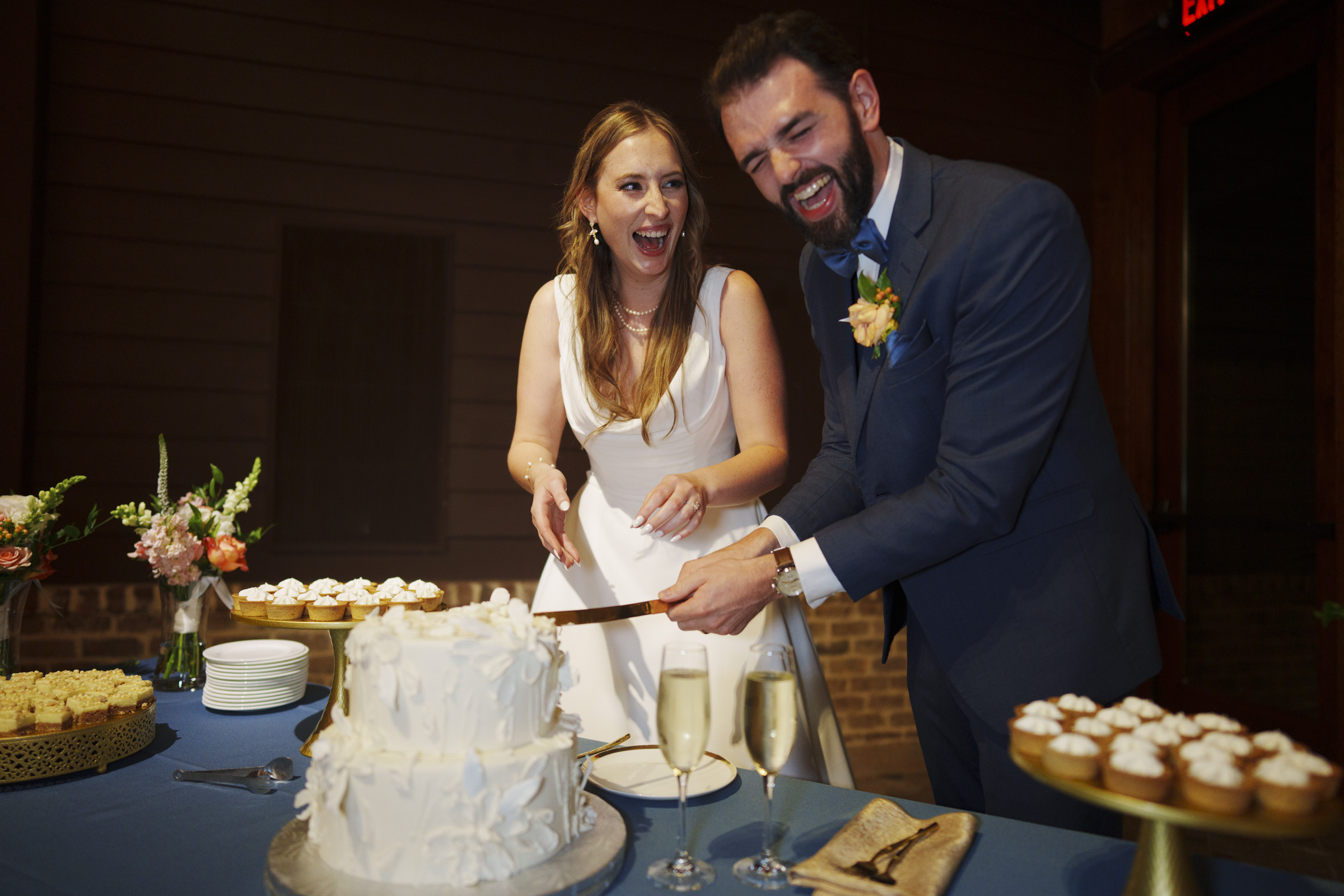 A bride and groom joyfully cut a white, three-tier wedding cake. They are both smiling widely. The table is adorned with champagne glasses and various desserts. The bride wears a white gown, and the groom is in a blue suit.