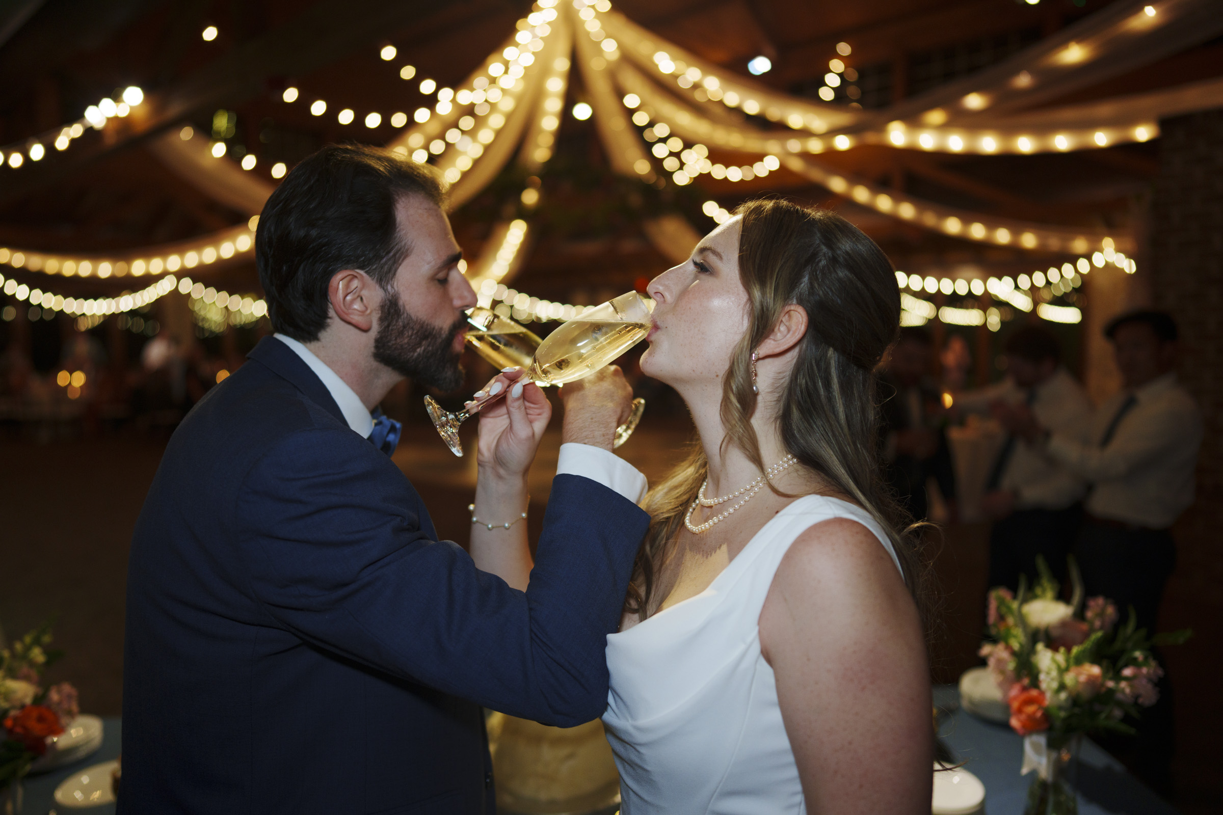 A bride and groom toast, interlocking arms while sipping champagne from glasses. Theyre under a canopy of string lights in a festively decorated venue. The bride wears a white dress, the groom a navy suit. Flowers adorn a nearby table.