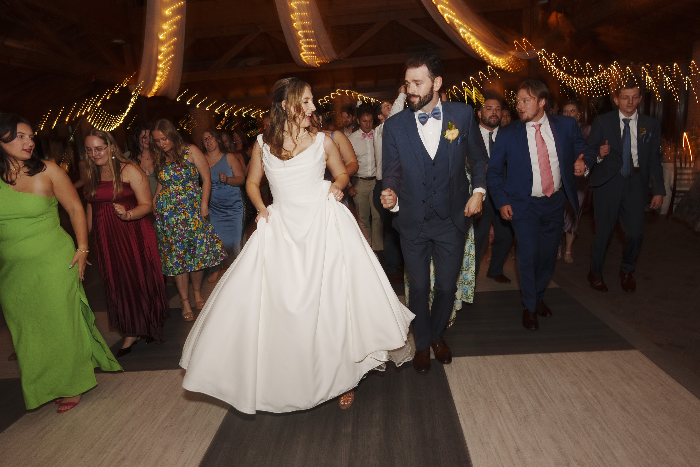 A bride in a white gown and a groom in a blue suit dance among a group of people at a wedding reception. The guests, dressed in colorful attire, are smiling and moving in unison under warm, string-lit decorations.