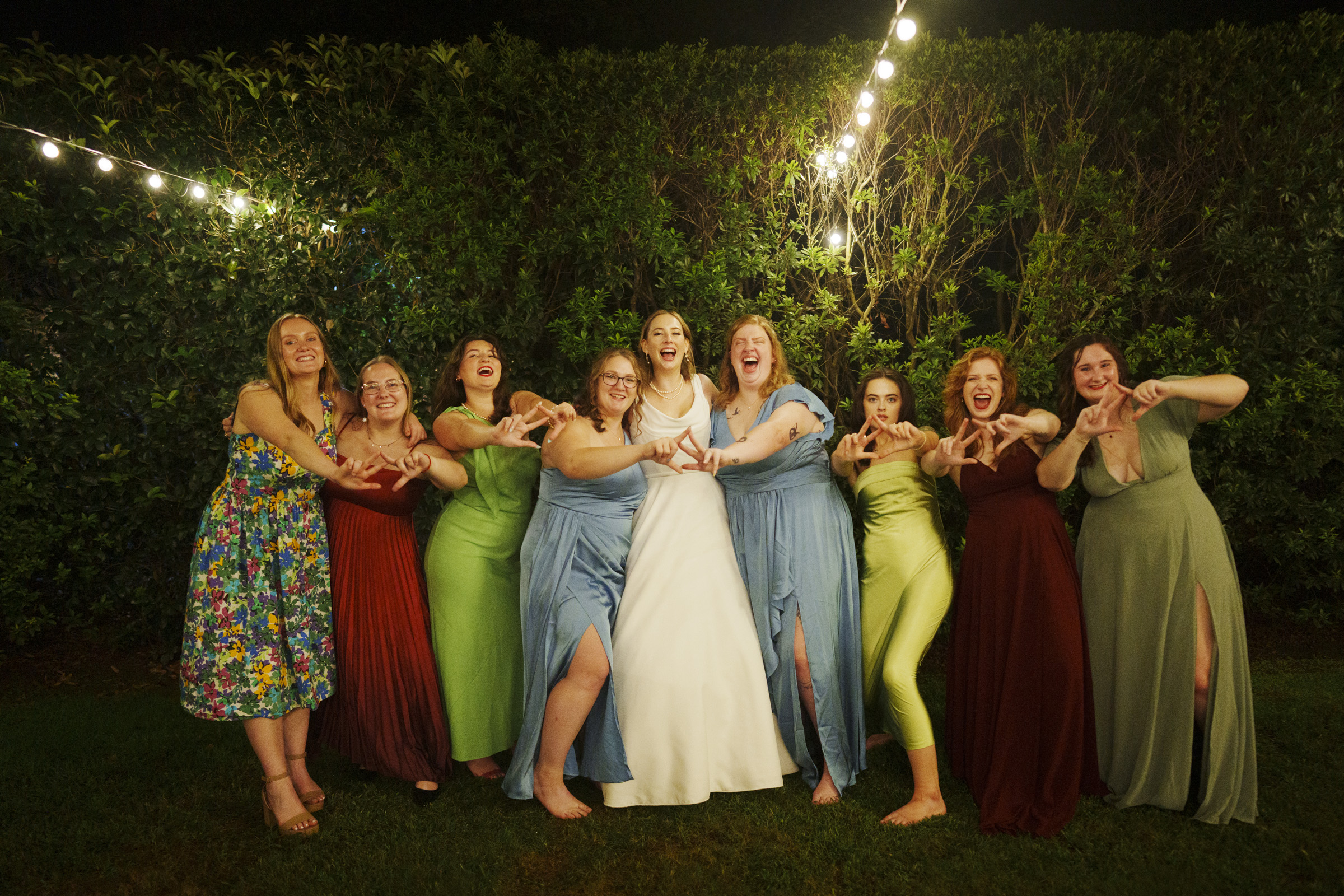 A bride in a white dress stands smiling with a group of bridesmaids and friends outdoors at night. The bridesmaids wear brightly colored dresses. They are posing joyfully with outstretched arms. String lights hang above, with trees in the background.