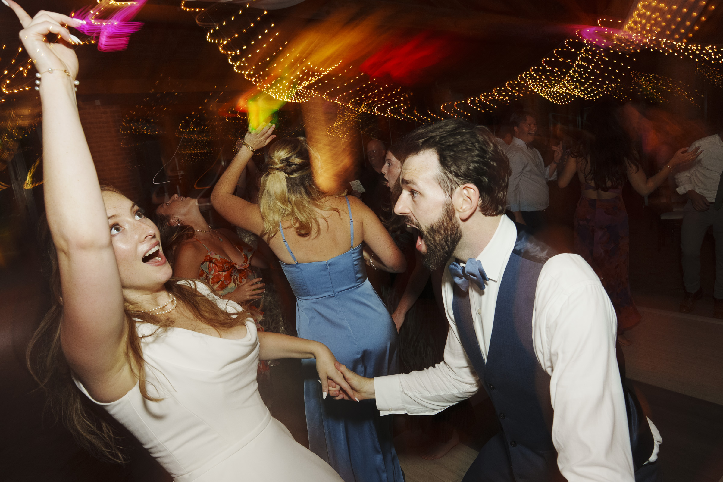 A bride in a white dress energetically dances with a bearded man in a blue vest and bow tie. Theyre surrounded by a lively crowd under strings of glowing lights, creating a festive atmosphere.