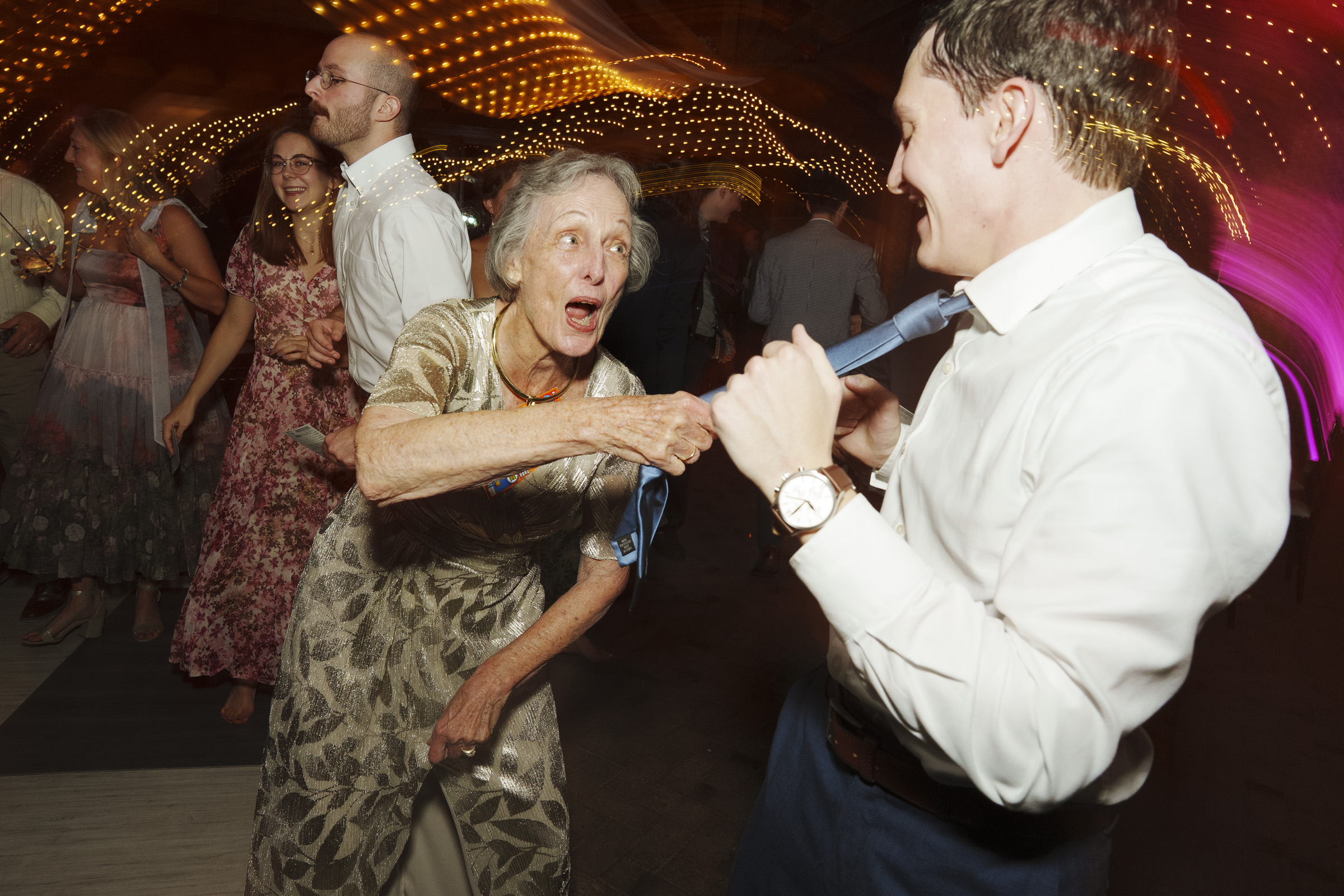 An elderly woman playfully pulls on a younger mans tie as they dance at a lively event. The woman is wearing a gold dress, and the man is in a white shirt. Blurred lights create a festive atmosphere in the background.