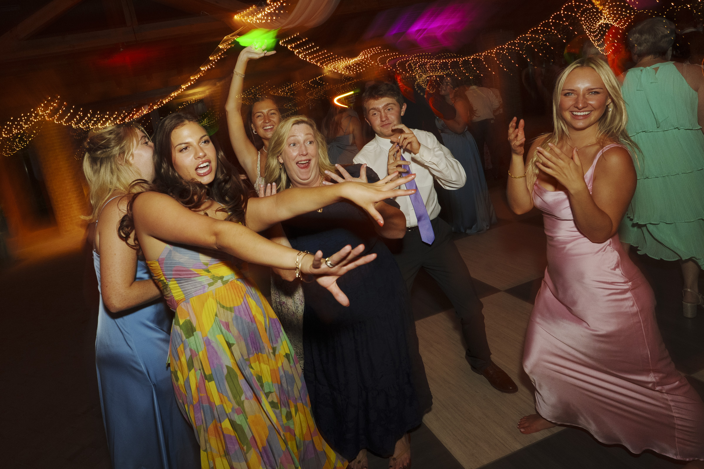 A group of people in formal attire dance energetically at an indoor party. They are smiling and posing under colorful lights with draped fabric and string lights in the background, creating a lively atmosphere.