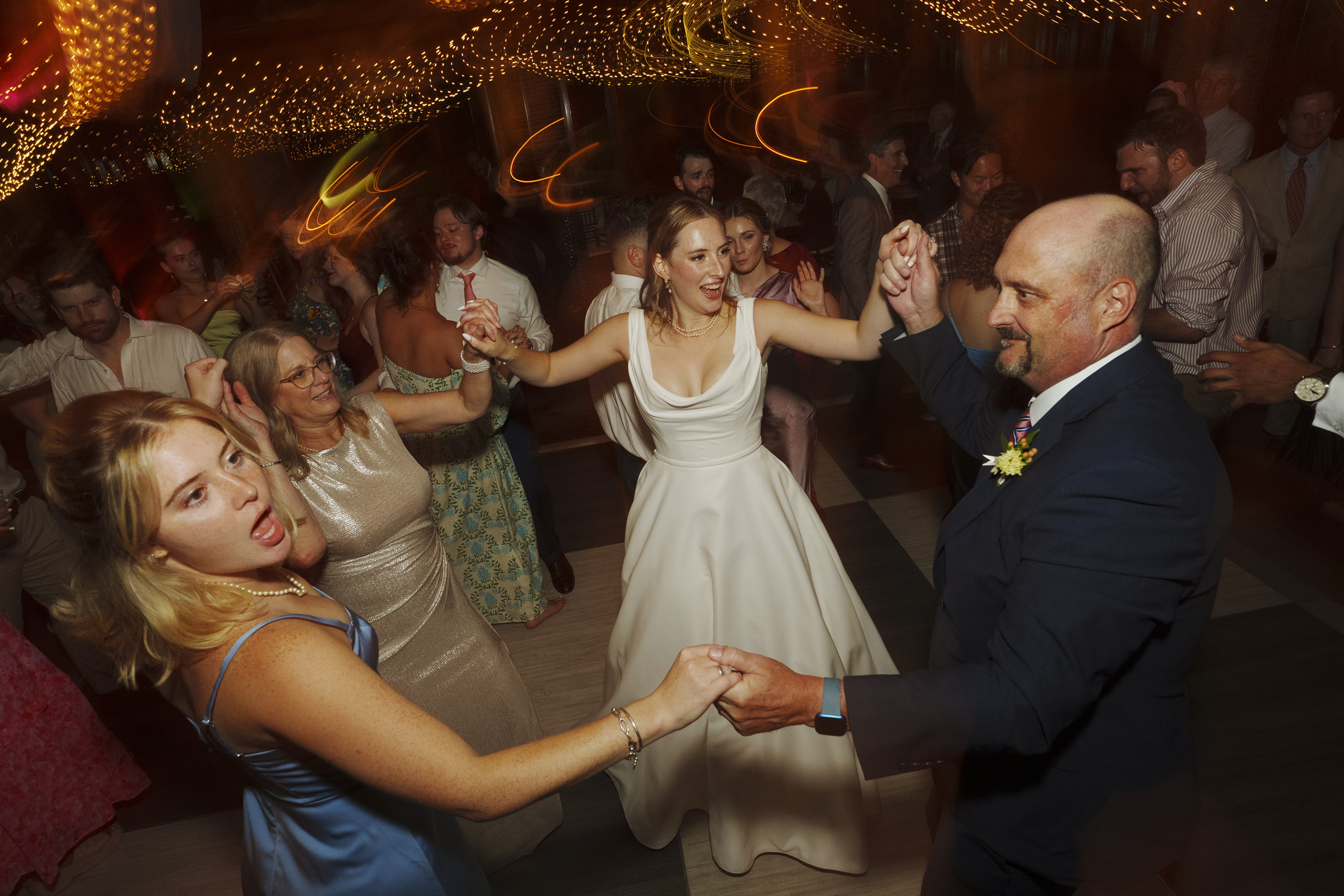 People are dancing joyfully in a dimly lit ballroom decorated with string lights. A bride in a white dress is at the center, surrounded by guests, holding hands in a circle. The atmosphere is lively and festive.