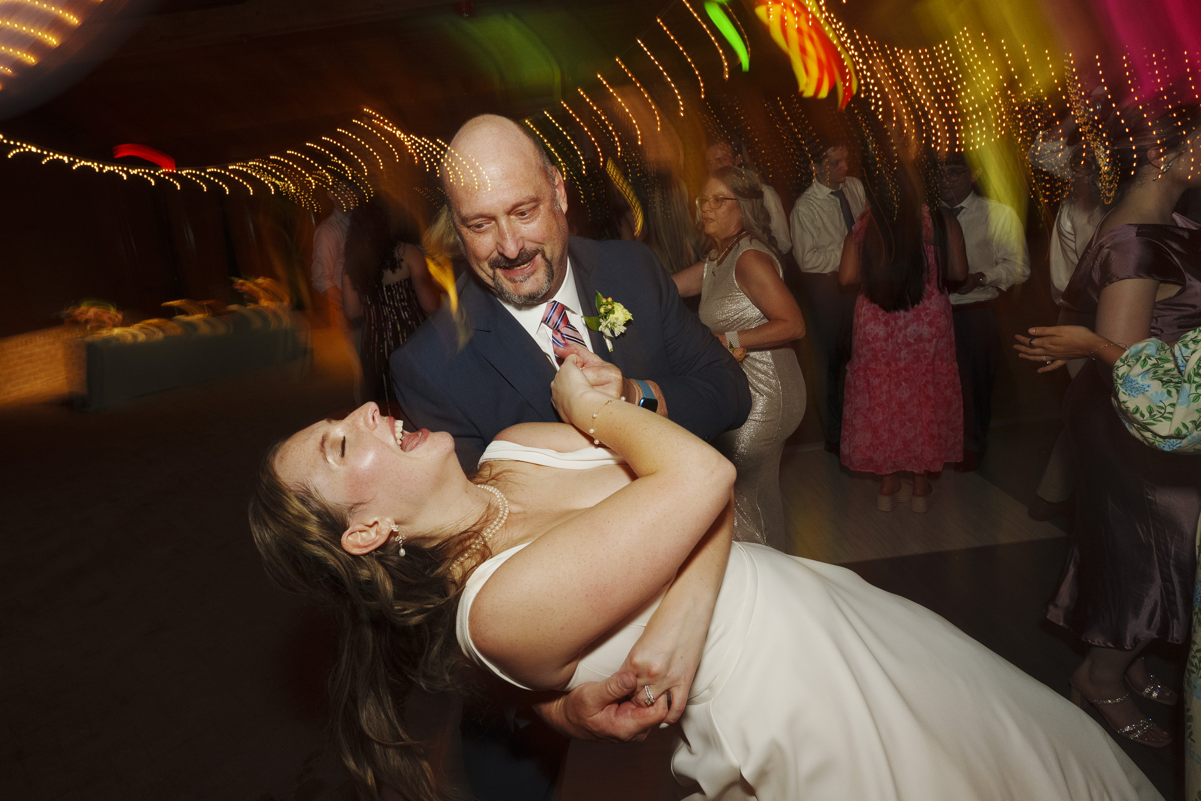 A bald man in a suit is dancing exuberantly with a woman in a white dress. She is laughing with her head tilted back. Other people in formal attire are dancing in the background with colorful lights overhead.