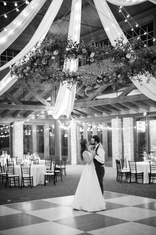 A couple dances alone on a wooden dance floor beneath a large, floral chandelier with draped fabric. The room is elegantly decorated with string lights, round tables, and dark chairs. The setting appears to be a wedding reception.