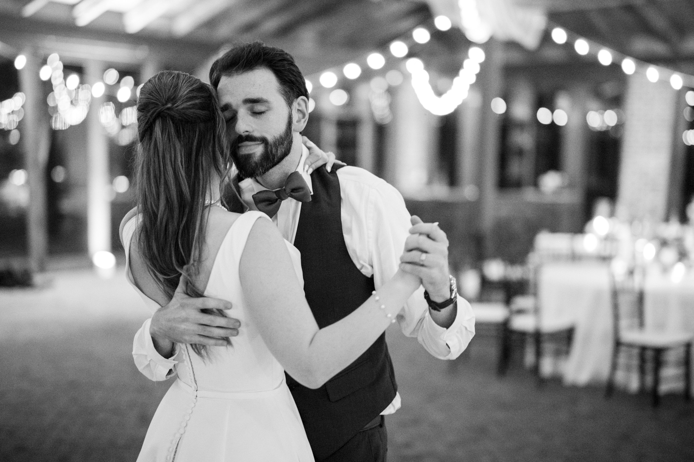 A couple dances closely in an elegant setting with string lights in the background. The man wears a vest and bow tie, while the woman is in a sleeveless dress. The atmosphere is intimate and warm.