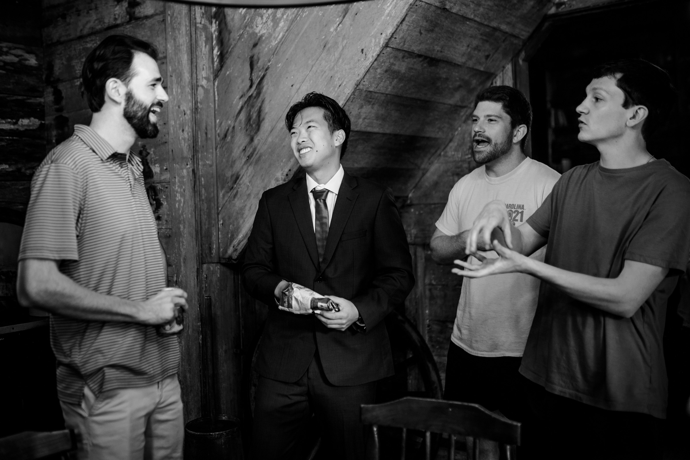 Four men are gathered in conversation indoors, with one wearing a suit and the others dressed casually. They appear engaged and cheerful, standing near wooden paneling and a chair in a dimly lit space.