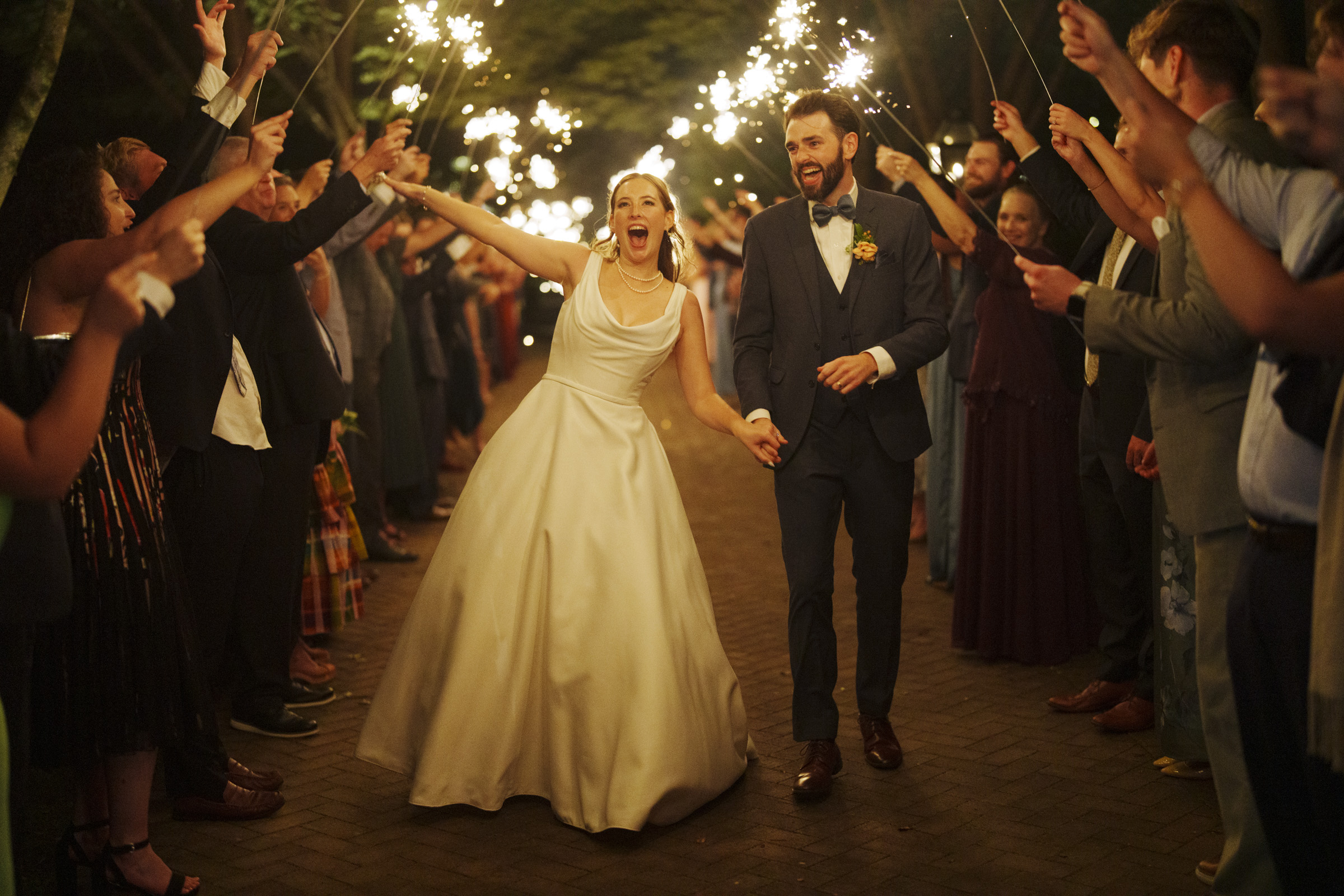 A joyful bride in a white dress and a groom in a suit walk through a pathway surrounded by guests holding sparklers. The couple appears excited, with the bride raising her arm enthusiastically. The scene is festive and vibrant.