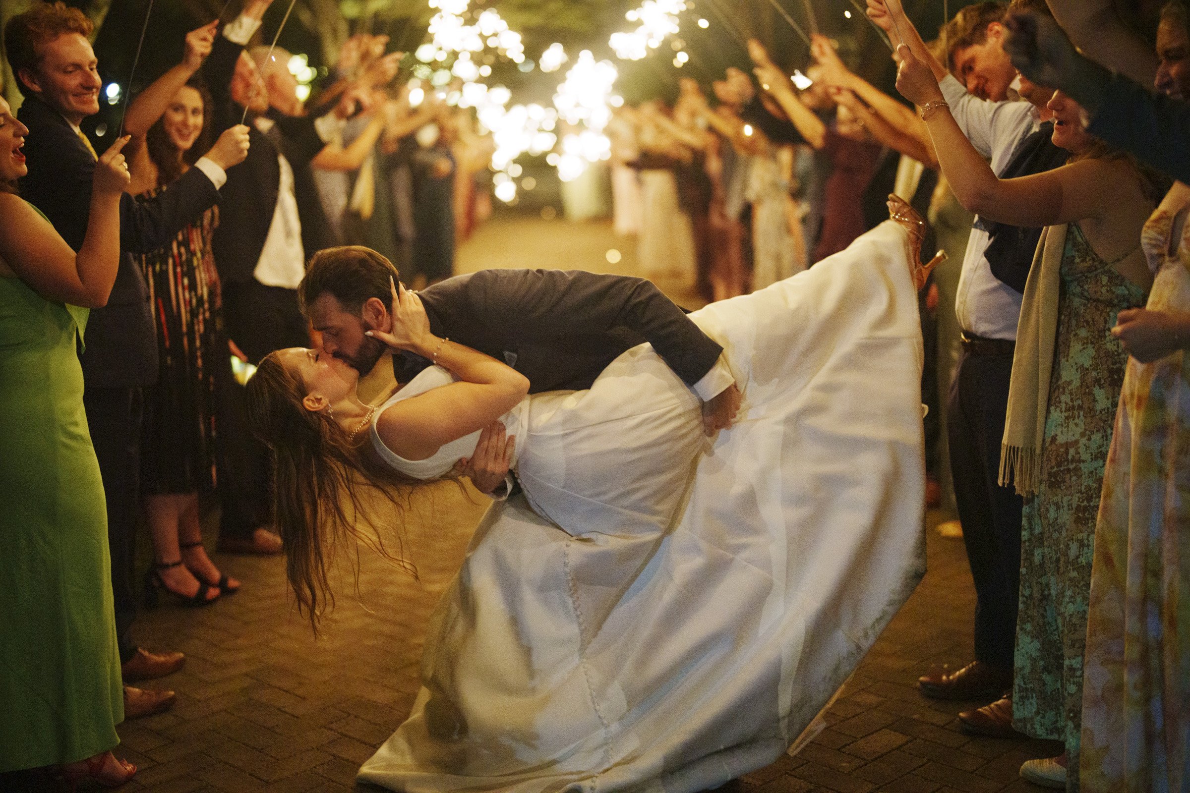 A newlywed couple shares a romantic kiss while the groom dips the bride in the center of a sparkler-lit path. Guests surround them in celebration, holding sparklers that illuminate the evening scene with warm, twinkling lights.