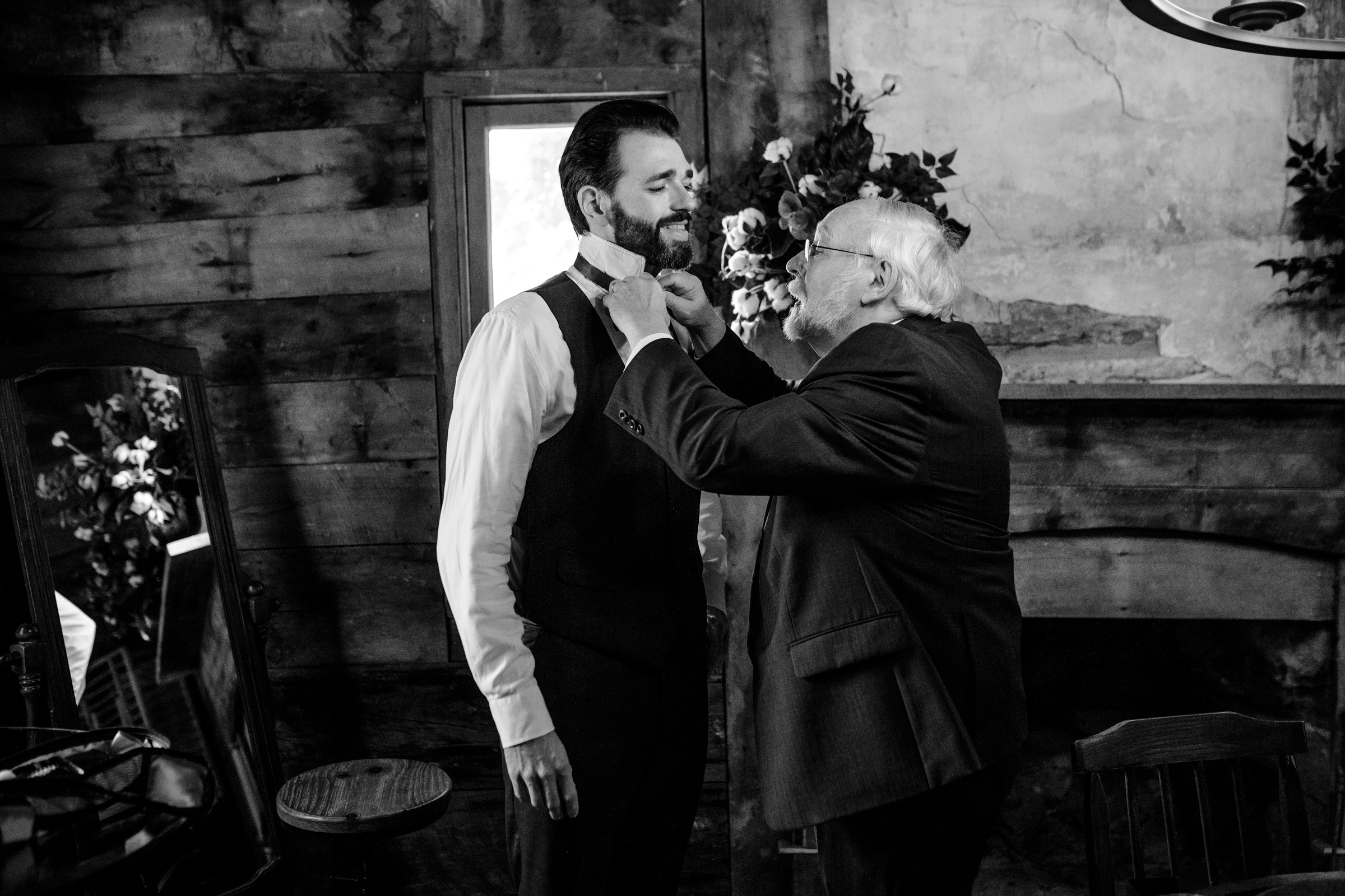 An older man adjusts the tie of a younger man in a suit, standing in a rustic room with wooden walls and floral decorations. A mirror and a chair are visible. The atmosphere is warm and intimate. The image is in black and white.